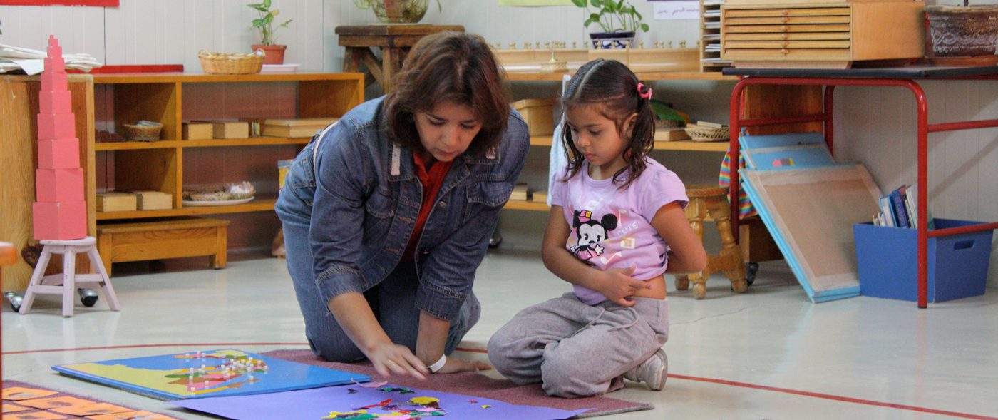 Guía Montessori acompañando a una niña que trabaja con un material en el piso del aula